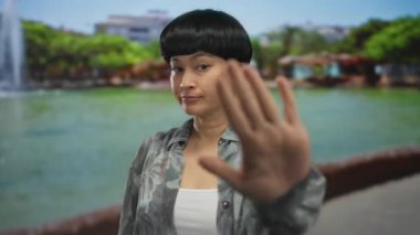 Arka planda yeşil ağaçlar olan açık hava parkında jest yapan bir kadın, su kenarında desenli gömlek giyen Çinli genç bir bayanı gösteriyor.