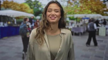 Kalabalık bir sokak pazarında arka planda satıcılar ve müşterilerle neşeli bir şekilde gülümseyen bir kadın, gün ışığında canlı bir açık hava sahnesini yakalıyor..