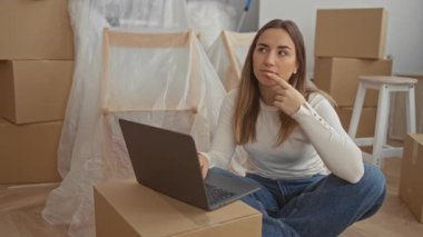 Karton kutularla çevrili yeni evde dizüstü bilgisayar kullanan bir kadın taşınma ve planlama fikirlerini yansıtıyor..