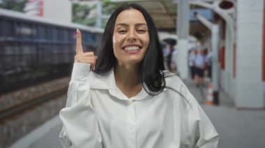 Beyaz gömlekli genç İspanyol kadın gülümsüyor ve açık hava tren istasyonunu işaret ediyor, bulanık demiryolu geçmişine karşı kurulmuş..