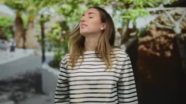 Güneşli bir parkta, açık havada gülümseyen genç bir kadın yemyeşil ve çizgili gömlekli..