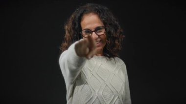 Kıvırcık saçlı ve gözlüklü gülümseyen kadın, elleri siyah arka planda bir kalbi işaret edip şekillendiriyor..