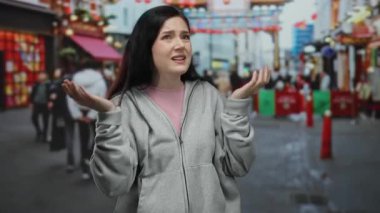 Sokakta kafası karışmış bir kadın, gri bir kazak giyiyor, renkli kentsel alanda dikiliyor insanlar ve canlı market tezgahları ile çevrili..