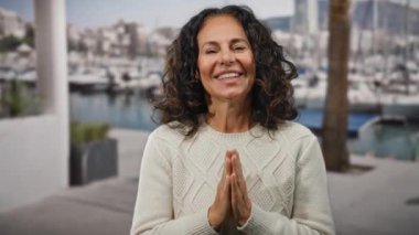 Sahildeki limanda gülümseyen bir kadın. Teknelerde kıvırcık saçlar sergileniyor ve beyaz bir süveter giyiyor. Suyun kenarında bulanık bir arka plan var..