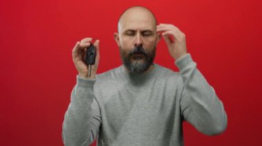 Sakallı, beyaz, kel bir adam arabanın anahtarlarını kırmızı arka planda tutarken sıkıntılı görünüyor. Gri bir süveter giyiyor, unutkanlık ya da kayıp hissi uyandırıyor..