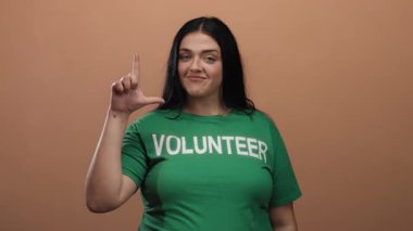 Yeşil gönüllü gömlekli kadın kahverengi duvara karşı ezik jestler yapıyor, mizah ve oyunculuk yapıyor..