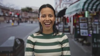 Neşeli bir sokakta gülümseyen mutlu kadın bulanık şehir geçmişiyle neşeli ifadesini ve günlük çizgili kıyafetini vurguluyor..