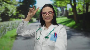 Parkta steteskopla gülümseyen İspanyol kadın doktor üniforma ve kırmızı gözlük takıyor, doğal ortamdaki bir sağlık hizmetleri profesyonel ortamını yakalıyor..