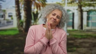 Gri saçlı yaşlı bir kadın, ağaçların altında pembe bir kazak giyerken parkta dua ediyor. Yüzünde huzur ve sükunet ifadesi var..