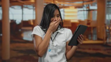 Oteldeki genç bir kadın tabletteki şaşırtıcı habere tepki veriyor, kapalı bir ortamda duygu ve haz ifade ediyor, eli ağzını şaşkınlık ve mutlulukla kaplıyor..