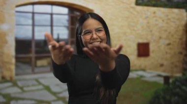 Güneşli bir günde kır evinin önünde kolları açık gülümseyen genç bir kadın. Gözlüklü ve siyah gömlekli. Mutluluğu iletiyor.