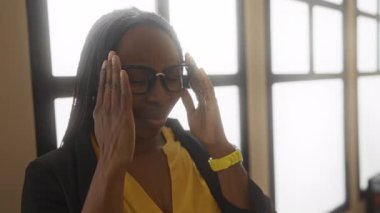Kıvırcık saçlı bir kadın ofis ortamında stresli görünüyor, gözlük takıyor ve sarı bir bluz giyiyor, hayal kırıklığı içinde kafasına dokunuyor..