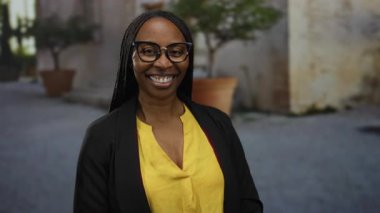 Sarı bir bluz ve gözlük takan bir kadın, bir şehir sokağında dikiliyor, mutluluğu ve kendine güveni iletiyor..