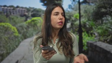 Güneşli bir günde Green Park 'ta akıllı telefon kullanan bir kadın, teknolojiye odaklanmış açık hava yaşam tarzını sergiliyor, genç bir kadın doğanın ortasında dijital iletişimle meşgul..