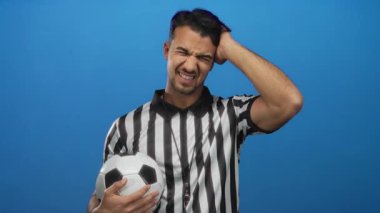 Çizgili tişörtlü İspanyol futbolcu hakem. İzole edilmiş mavi arka plan üzerinde düşünceli bir ifadeyle topu tutuyor..