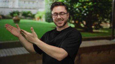 Parkta sakallı ve gözlüklü genç adam, güneşli bir günde yeşil arka planda bir şey sunan, neşeli bir yüz ifadesi gösteren elini uzatıyor..