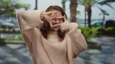 Parkta bir kadın fotoğraf jesti yapıyor, gözlük ve süveter takıyor, açık havada ağaçlarla ve palmiyelerle gülümsüyor, doğal olarak yakalanan sıradan ve neşeli bir anı işaret ediyor..