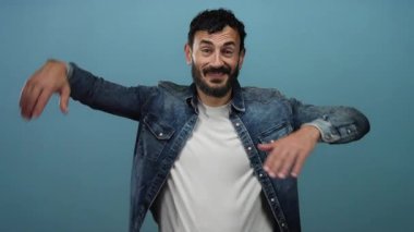 Sakallı İspanyol bir adam mavi arka planda şakacı poz veriyor, etkileyici hareketleri neşeli ve kendinden emin bir tavır sergiliyor..