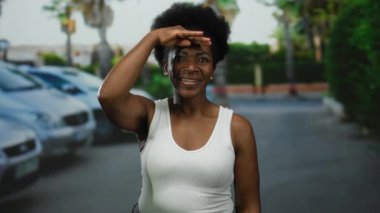 Dışarıda gülümseyen bir kadın, güneşli şehir caddesinde el gölgesi gibi gözleri olan, arka planda arabalar ve ağaçlarla, kentsel çevreye pozitif ve güven yansıtan bir kadın..