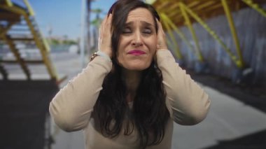 Kadın stresli görünüyor, şehir caddesinde kulaklarını kapatıyor, açık hava ortamında duygularını ifade ediyor, binaların arka planı ve açık mavi gökyüzü ile.