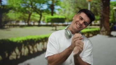 Kulaklıklı genç adam, yeşil ağaçlar ve patikalarla çevrili güneşli bir parkta gülümsüyor, rahat bir açık hava havasını yansıtıyor ve gündelik bir stili yansıtıyor..