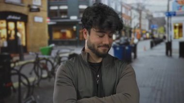 Sakallı ve çapraz kollu bir İspanyol genç. Bulanık dükkanları ve bisikletleriyle şehir sokağında güvenle duruyor. Açık havada moda tarzını sergiliyor..