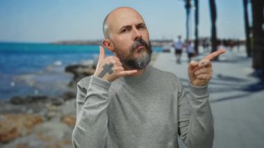 Bald bearded man gesturing the phone symbol on a seaside promenade with the ocean in the background demonstrating a relaxed outdoor scene.