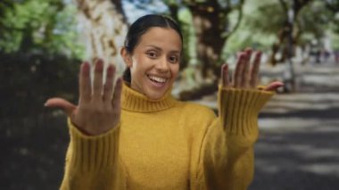 Parkta gülümseyen sarı bir kazak giyen bir kadın yeşil ağaçların arasında elleri ile bir hoş geldin jesti yapıyor. Açık havada sıcaklığı ve ulaşılabilirliği gösteriyor..