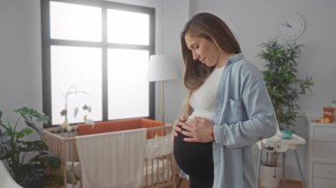 Sarışın hamile bir kadın göbeğini, yumuşak ortam ışığı altında bir beşik, bitkiler ve yüksek bir sandalyeyle çevrili huzurlu bir bebek odasında sevgiyle kucaklıyor..