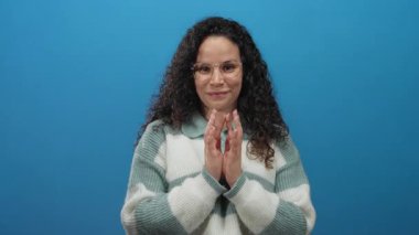 Kıvırcık saçlı, elleri sımsıkı bağlı bir kadın, parlak mavi bir arka plan karşısında minnet ve samimiyetini gösteriyor..