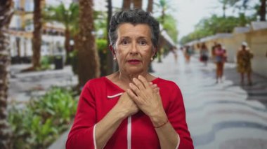 Meşgul şehir caddesinde kırmızı kazaklı yaşlı bir kadın çerçevenin dışında bir şeyden ürkmüş, palmiye ağaçları ve arka plandaki insanlarla şehir yaşamını gözler önüne seriyor..