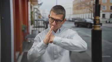 Savunma jesti olarak gözlüklü beyaz bir adam sakin bir sokakta açık şehir ortamında duruyor, hazır olduğunu ve kararlılığını gösteriyor.