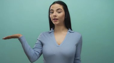 Mavi duvara karşı el ve baş parmaklarıyla el kol hareketi yapan, mavi bluz giyen, canlı bir iç mekanda pozitiflik ve iletişim kuran bir kadın..