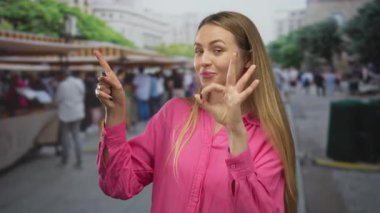 Pembe tişörtlü bir kadın şehir pazarında gülümseyip işaret ederken insanların ve tezgahların bulanık arkaplanlarını gösteriyor..