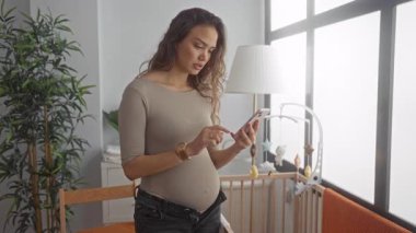Bebek beşiği ve yeşillikle parlak bir yatak odasında telefon kullanan hamile bir kadın, huzurlu ve modern bir anne atmosferi yaratıyor..