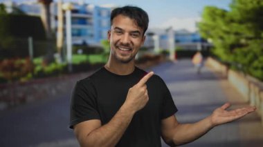 Siyah tişörtlü genç İspanyol adam gülümsüyor ve modern şehir geçmişine sahip güneşli bir caddede işaret ediyor..