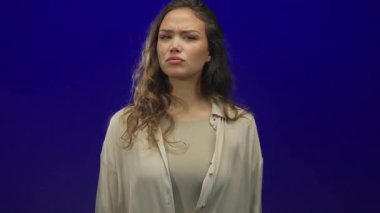 Latin kökenli genç bir kadın, mavi stüdyo ortamında el hareketini durdurarak el kaldırıyor..