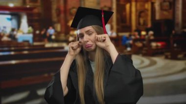 Mezuniyet kıyafetli bir kadın, arka planda bulanık iç kısımları olan büyük bir kilisenin içinde hüzün ile alay ederek mizahi bir jesti vurguluyor..