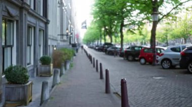 Arka planda bokeh efektli bir Amsterdam caddesinin bulanık perspektifi, ağaç kaplı kaldırımlar, park edilmiş arabalar ve tarihi mimari sergileniyor..