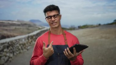 Gözlüklü ve kırmızı kazaklı genç bir adam, elinde tablet, sakin bir caddede gülümsüyor..