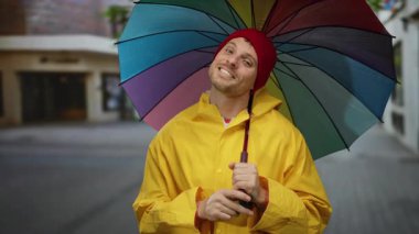 Sarı yağmurluklu adam şehir caddesinde renkli bir şemsiye tutuyor.