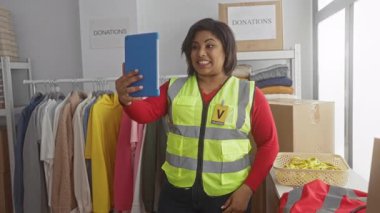 Kıyafet ve kutularla dolu bir bağış merkezinden yansıtıcı yelek giymiş bir kadın arıyor. Canlı bir toplum gönüllüsünü kapalı alanda sergiliyor..