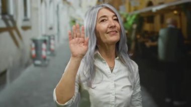 Gri saçlı, gülümseyen ve canlı bir şehir sokağında el sallayan bir kadın şehir ortamında kendine güvenini ve sıcaklığını gösteriyor..
