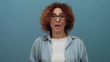 Orta yaşlı İspanyol kadın, kısa kıvırcık esmer saçlı, gözlüklü ve günlük kıyafetli, izole edilmiş mavi arka plana şaşırdığını ifade ediyor..