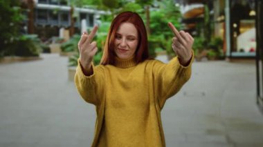 Şehir sokağında orta parmaklı bir kadın, sarı bir kazak giyiyor, kızıl saçlı, kendine güvenen bir ifade çiziyor, şehir arka planında bulanıklık var, davranışlarını vurguluyor.