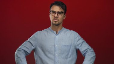 Gözlüklü ve mavi tişörtlü İspanyol genç adam avucunun içine parlak kırmızı arka plana yumruk atıyor, izole stüdyo ortamında öfke ve kararlılık taşıyor..