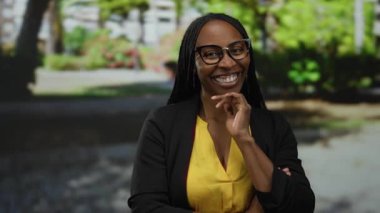 Açık havada, yemyeşil bir parkta, sarı bluz ve siyah ceket giymiş, canlı kıvırcık saçlar ve şık gözlükler takan bir kadın..