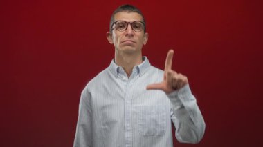 Erkek, kırmızı arka plandaki izole edilmiş kırmızı duvara işaret ederken ezik işareti yapar, çizgili gömlek ve gözlük giyer, onaylamadığını belirtir ve odaklanır..