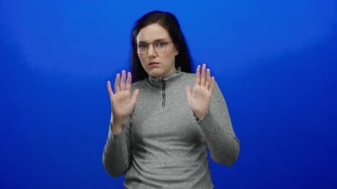 Mavi arka planda gözlüklü ve gri kazaklı, şaşırmış ve anlamlı görünen bir kadın..