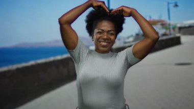 Deniz kenarında gülümseyen bir kadın, güneşli mavi gökyüzünün altında kalp hareketi yapar..